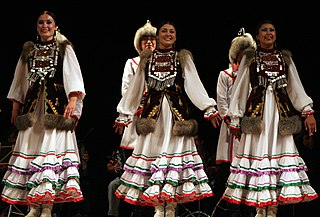 Tres bailarinas en un escenario. Llevan vestidos largos blancos con tres volantes adornados en diferentes colores. Sobre los vestidos llevan chalecos de color marrón oscuro cubiertos con discos plateados y ribeteados de piel. Al fondo se ven dos hombres con gorros de piel.