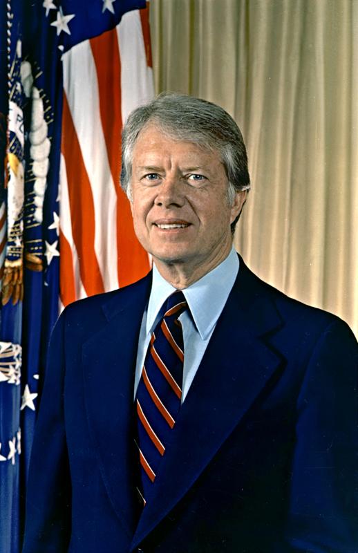 Picture of Jimmy Carter. He is wearing a blue suit, in front of yellow backdrop. There are American flags beside him. He has medium hair and no facial hair and is smiling. 