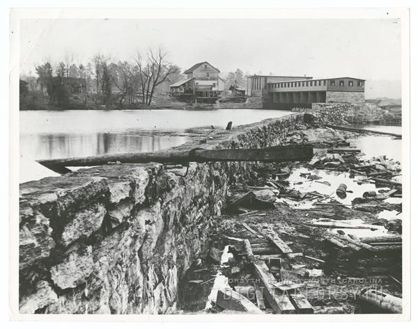 Idol's dam. A reservoir of water is held by a stone wall. A facility sits at the top of the reservoir next to a home. Trees surround the reservoir. 